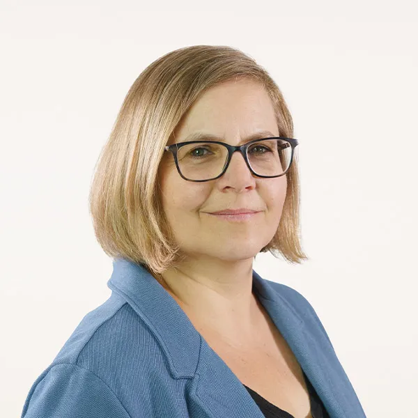 Porträtfoto von Marion Elle, Ansprechpartnerin des Projektes, mit blonden Haaren, Brille und blauem Blazer vor hellem Hintergrund.